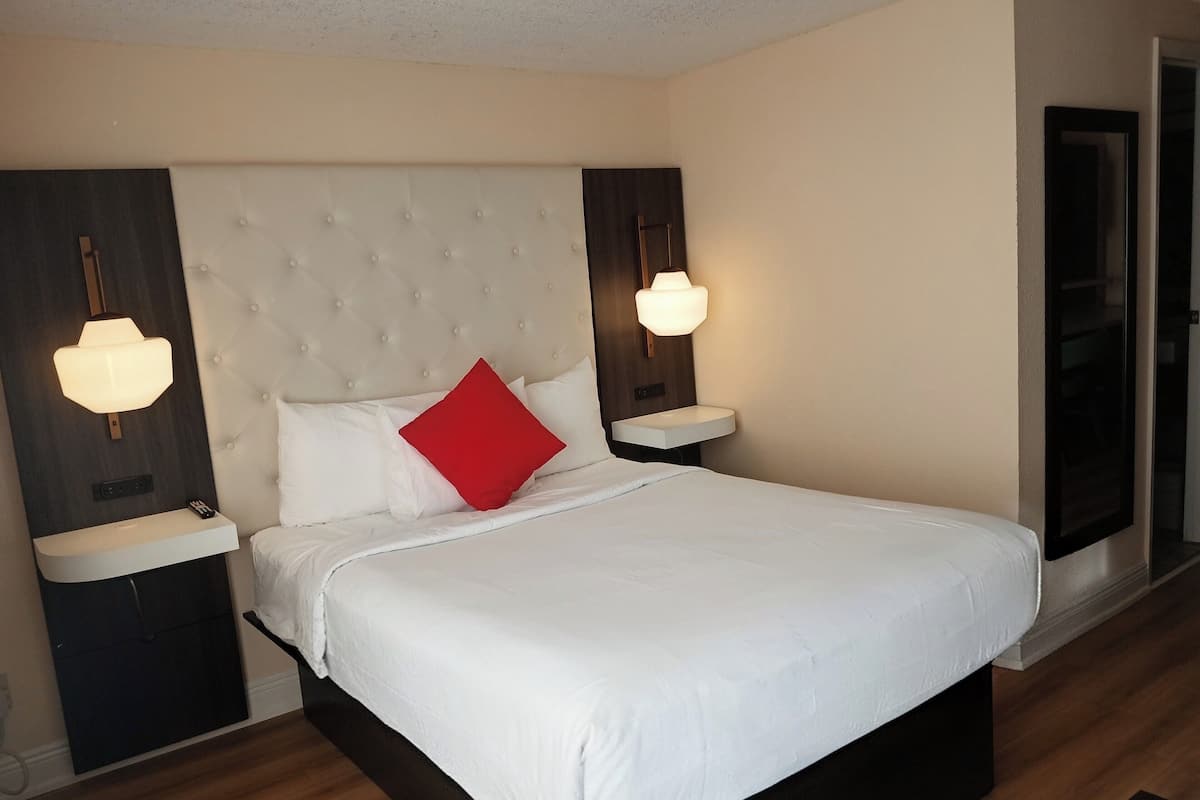 A hotel room with a large bed, a bedside table, a nightstand, a wall-mounted light fixture, and a large window.