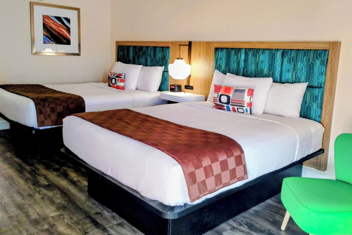 A hotel room with two beds, a green chair, a nightstand with a lamp, and a framed picture on the wall.
