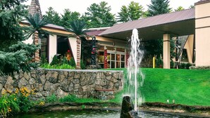 A fountain in a landscaped area with a stone wall, a building with a sloped roof, and trees in the background. - Tiki Resort (Lake George)
