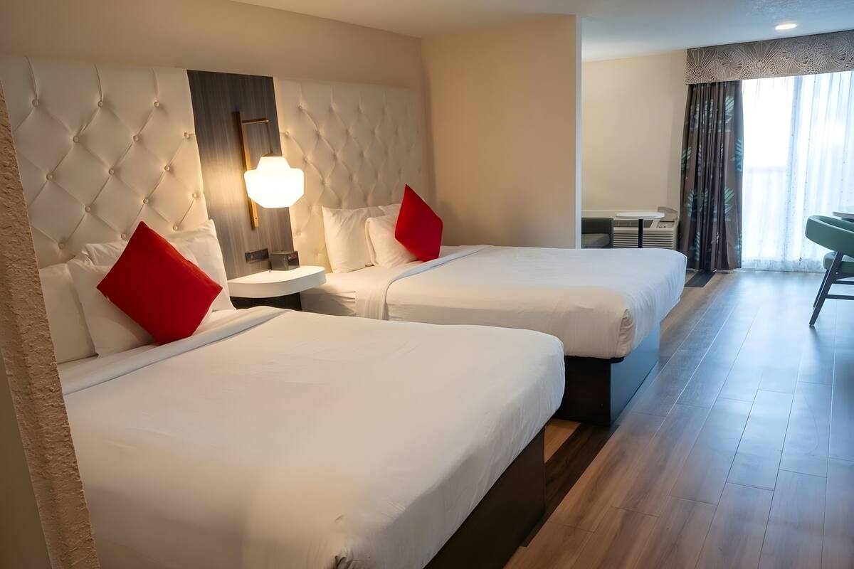 A hotel room with two beds, a nightstand, a lamp, and a view of the outside through a window.