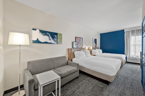 Une chambre d’hôtel avec un canapé gris, deux lits simples, une petite table et une lampe sur pied.