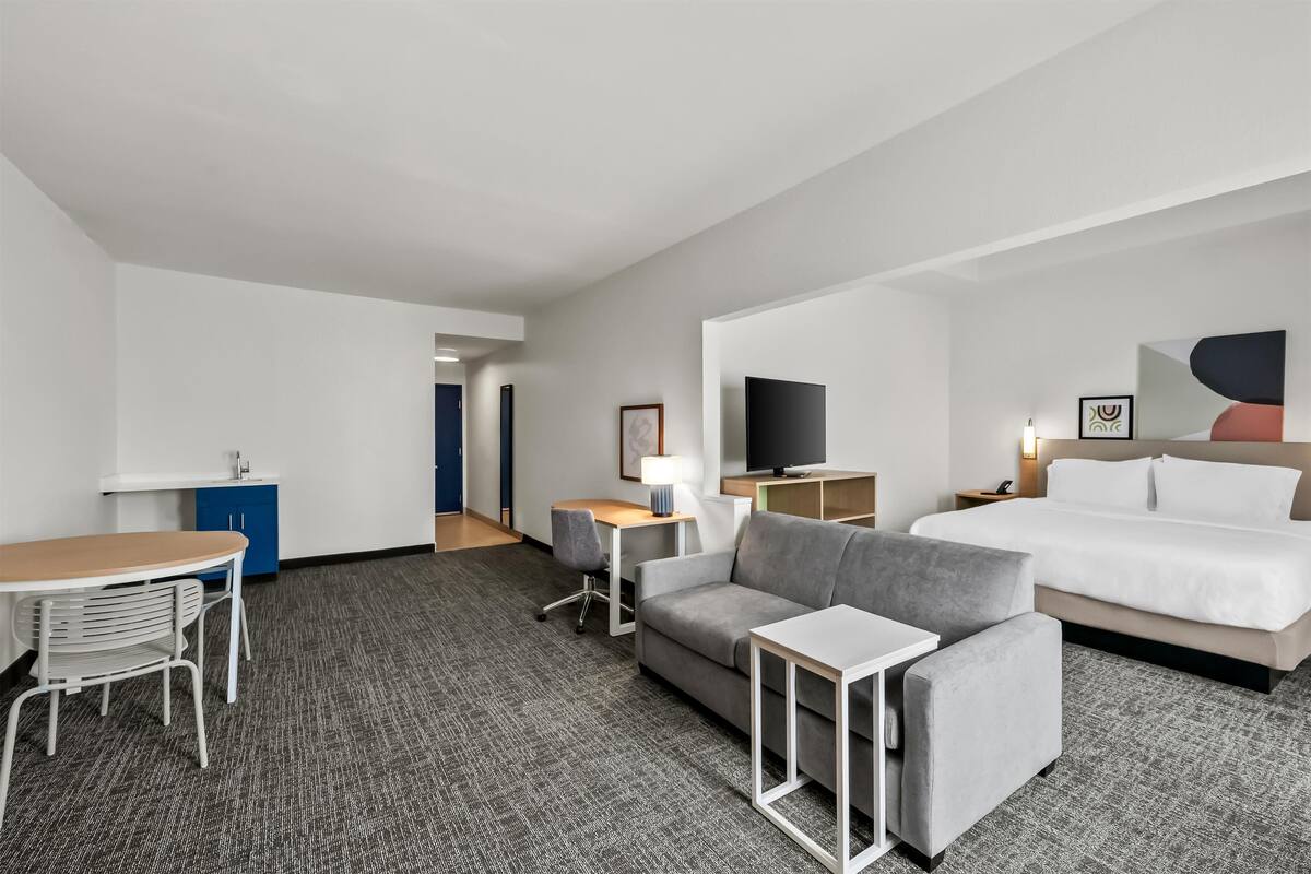 Une chambre d’hôtel comprenant un lit, un canapé, un bureau avec une lampe, une télévision et une petite kitchenette.