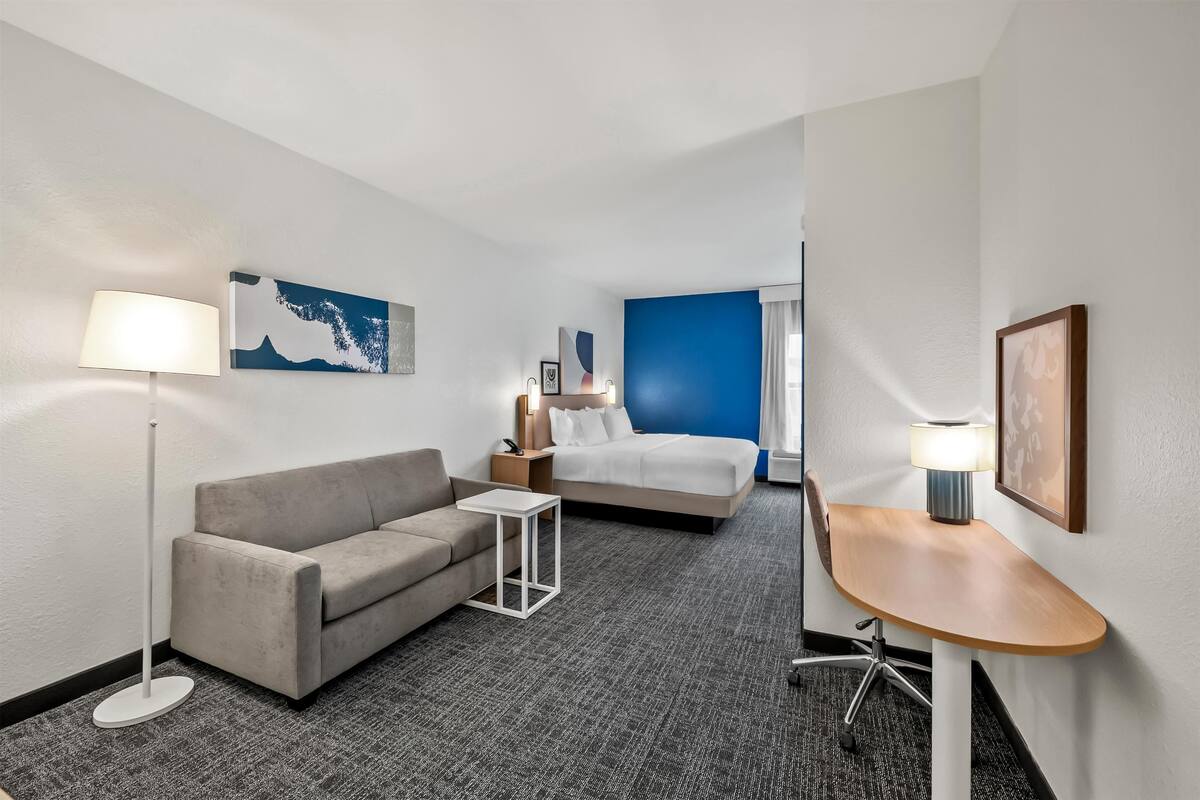 Une chambre d’hôtel avec un canapé gris, une petite table blanche, un lit avec un mur d’accent bleu et une table de chevet en bois.