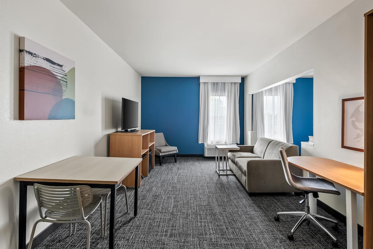 Une chambre d’hôtel équipée d’un bureau, de deux chaises, d’un canapé, d’une télévision et d’une grande fenêtre avec des rideaux.