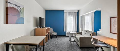 Habitación de hotel con escritorio, dos sillas, sofá, televisor y un amplio ventanal con cortinas.