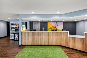 A hotel reception area with a wooden reception desk, a green carpet, and a wall with a colorful abstract design.