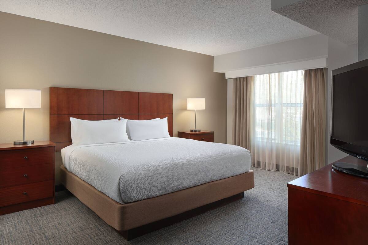 A hotel room with a large bed, two bedside tables with lamps, a flat-screen TV, and a large window with curtains.