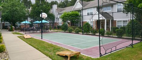 A residential outdoor tennis court with a wooden bench, a basketball hoop, and a covered seating area.