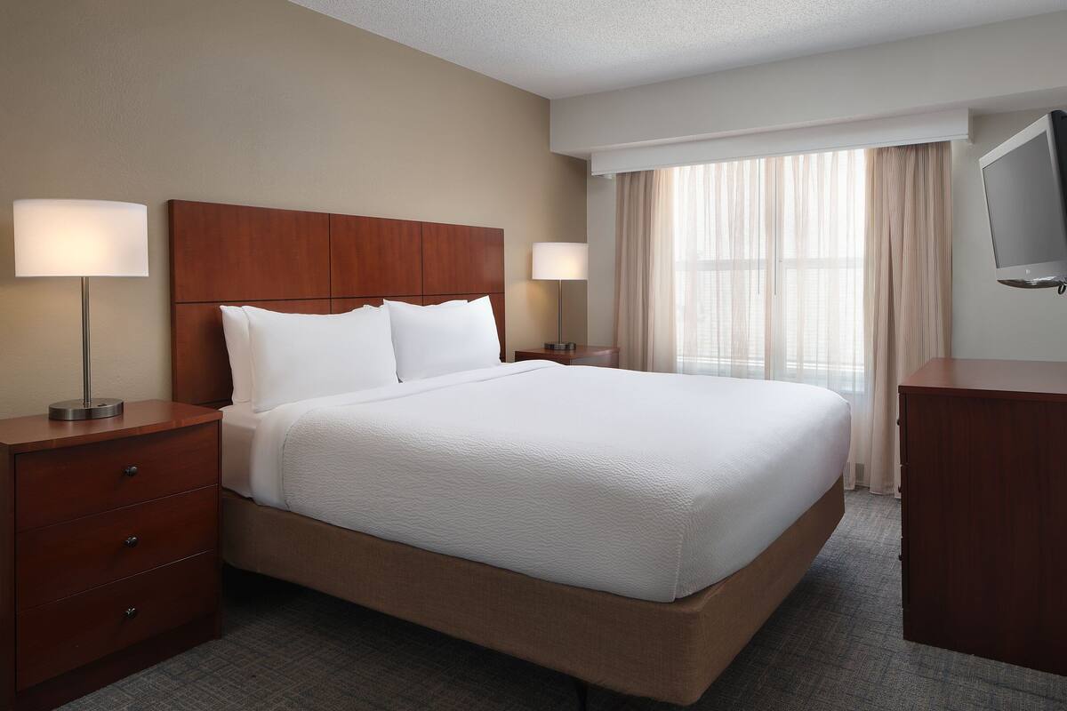 A hotel room with a large bed, two bedside tables with lamps, a flat-screen TV, and a window with curtains.
