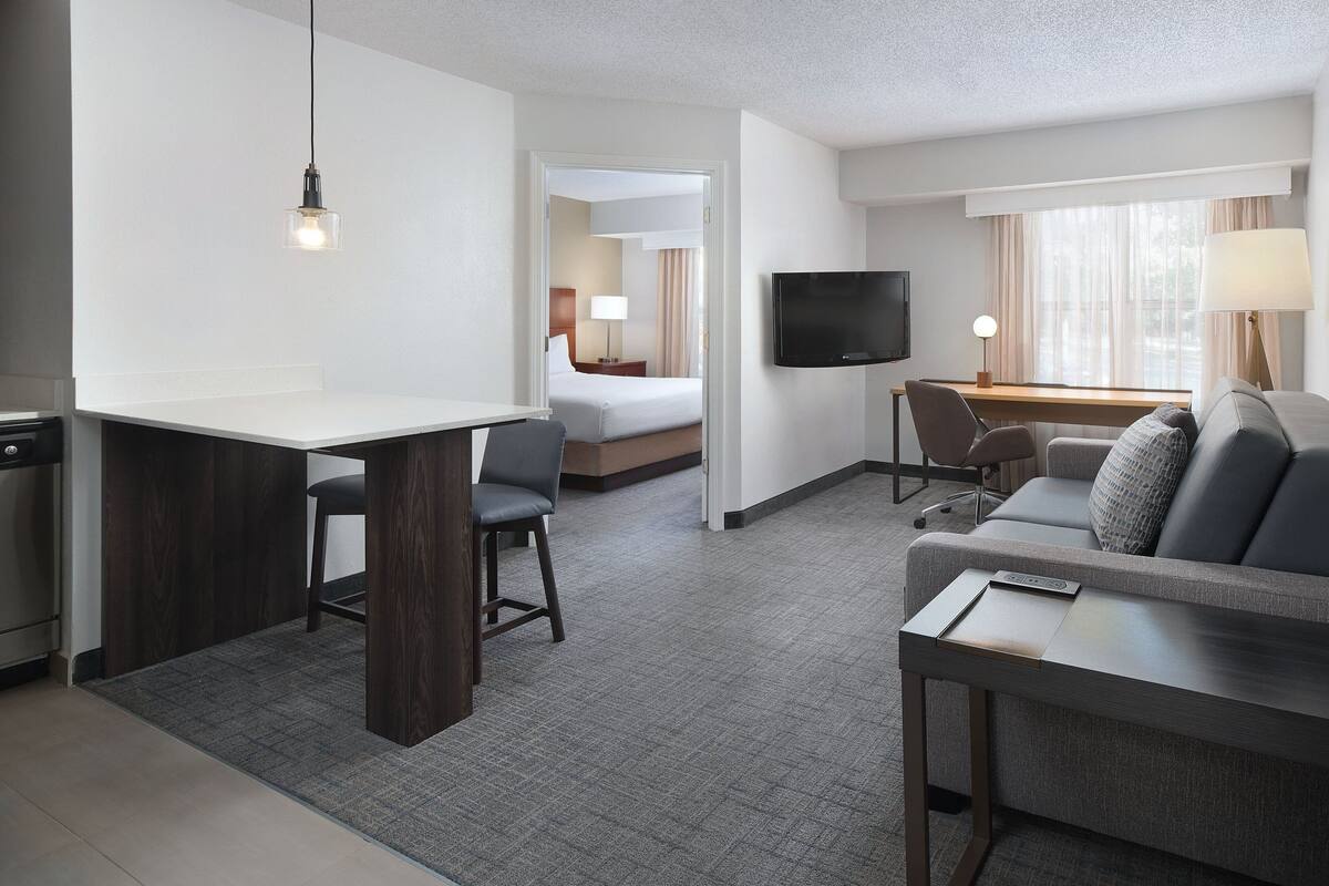 A hotel room with a kitchenette, a dining area, a living area with a sofa and TV, and a separate bedroom with a bed and window.