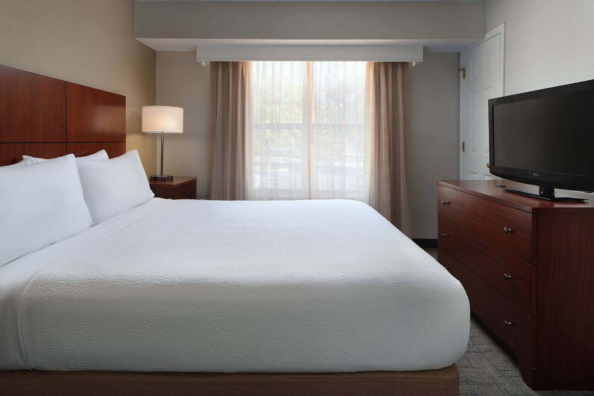 A hotel room with a large bed, a TV on a wooden dresser, and a window with curtains.