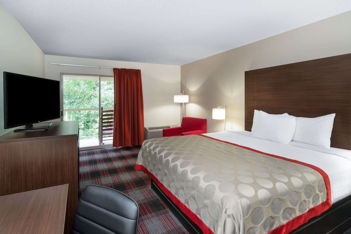 Une chambre d’hôtel avec un lit, un bureau avec une télévision, une chaise rouge et une grande fenêtre avec des rideaux.