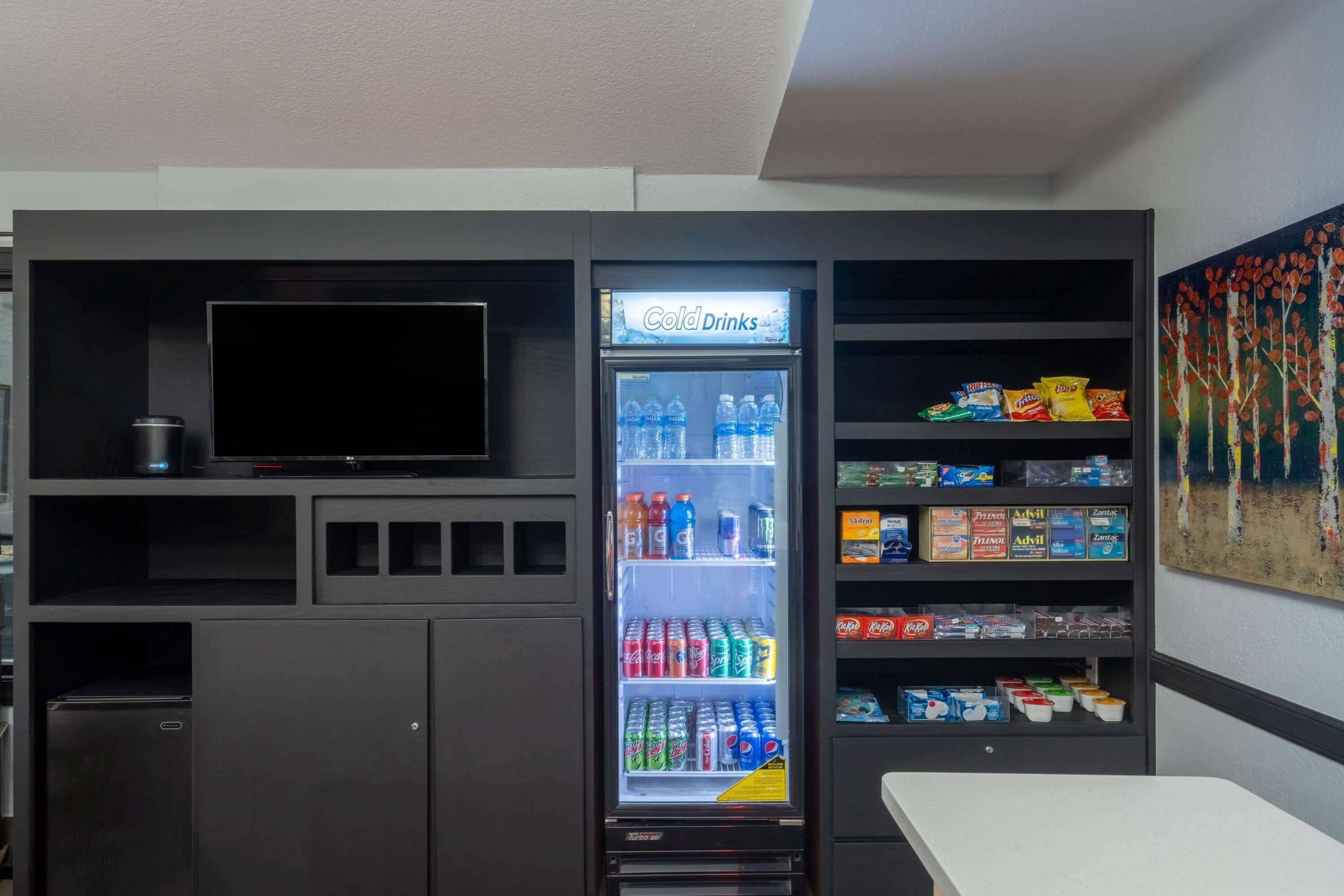 Een automaat met diverse drankjes en snacks, een televisie bevestigd aan een donkere kast, en een schilderij aan de muur.