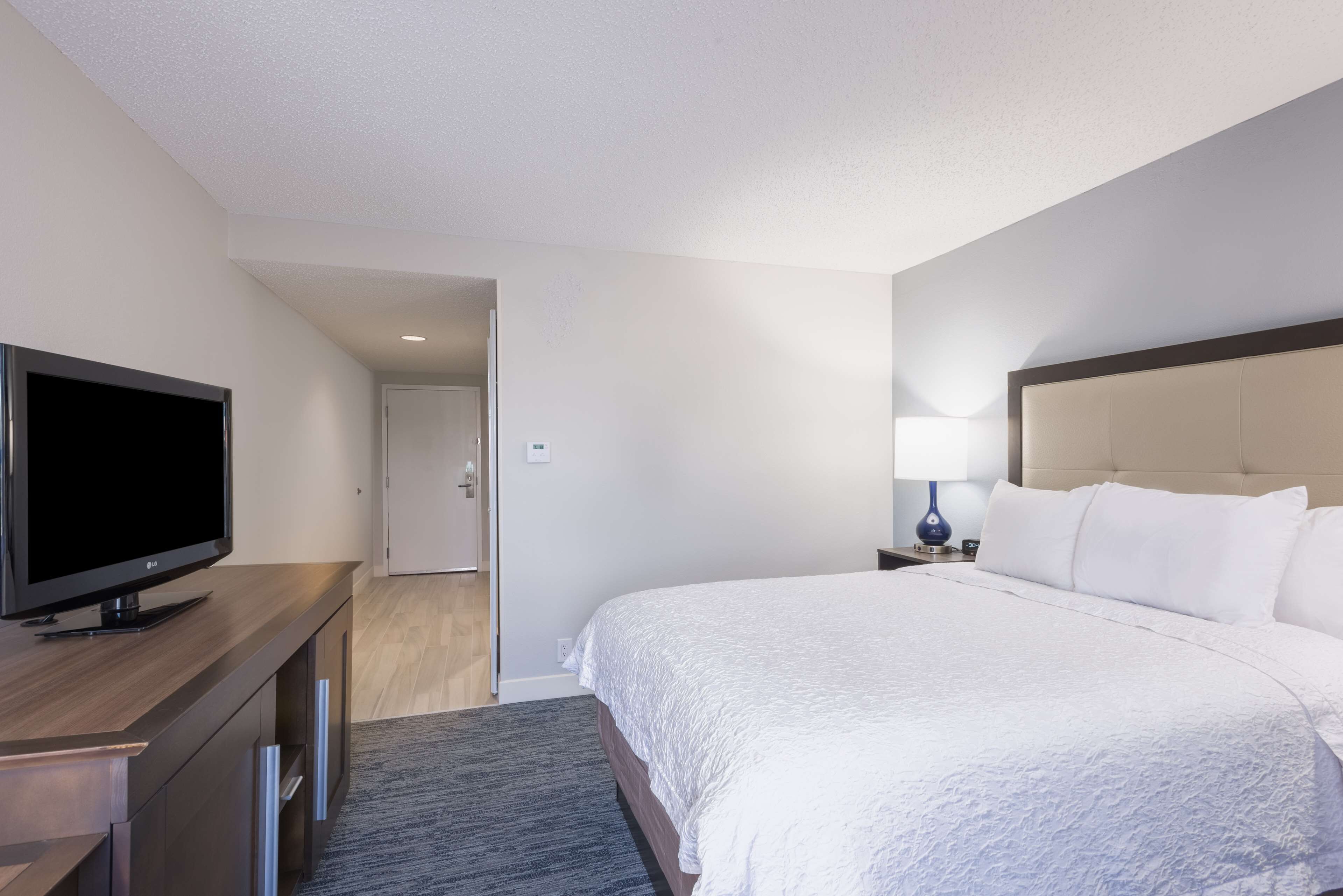 A hotel room with a bed, a television on a wooden dresser, a nightstand with a lamp, and a door leading to another room.