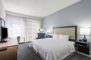 Habitación de hotel con una cama grande, un escritorio con televisor, una silla, una mesita de noche con lámpara y una ventana con cortinas.