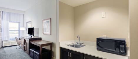 A hotel room with a kitchenette, a desk with a chair, a television, and a window with curtains.