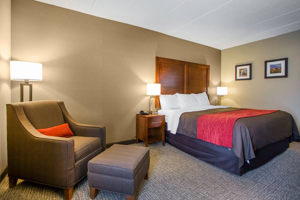 A hotel room with a bed, a chair, an ottoman, two bedside lamps, and a nightstand.
