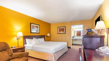 A hotel room with a large bed, a desk with a lamp, a chair, and a small table with a telephone and papers.