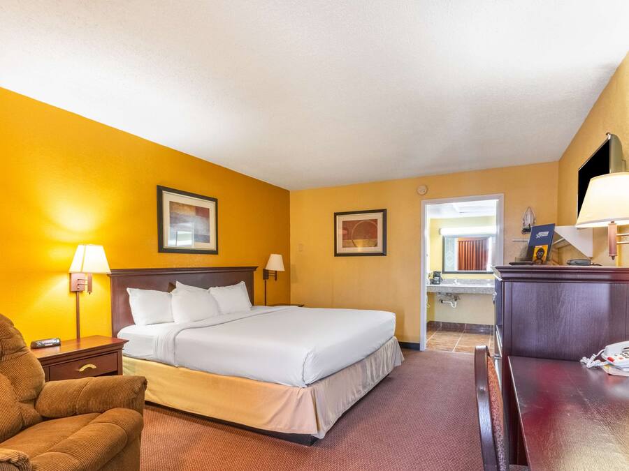 A hotel room with a large bed, a desk with a lamp, a chair, and a small table with a telephone and papers.