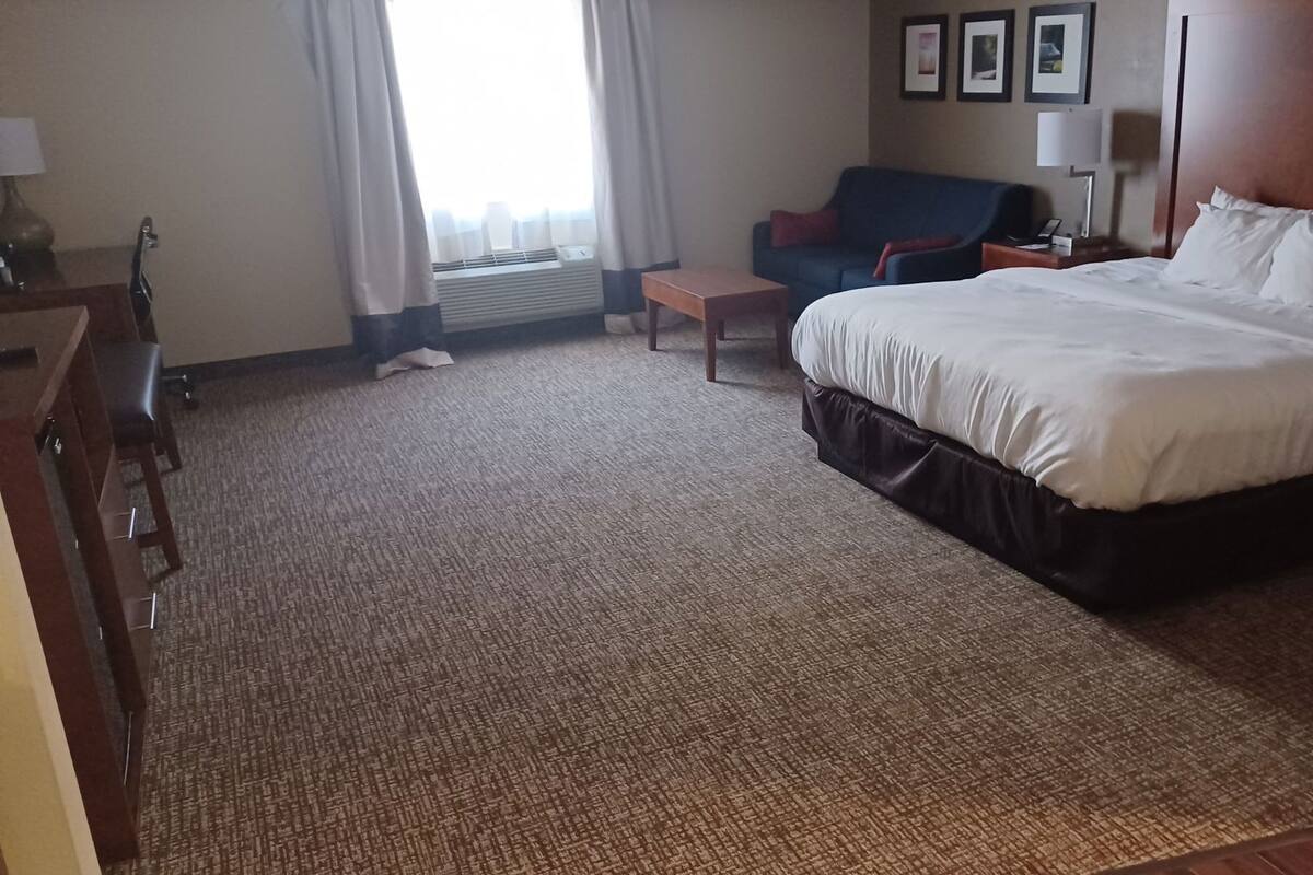 Et hotelværelse med en seng, et skrivebord, en stol, en sofa, et lille bord og et vindue med gardiner.