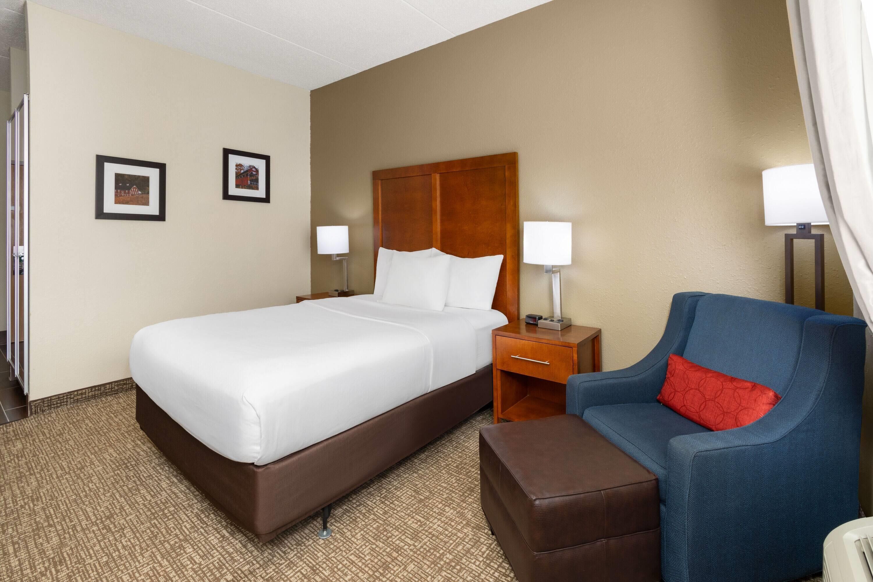 A hotel room with a large bed, a wooden headboard, two bedside tables with lamps, a blue armchair with a red pillow, and a brown ottoman.