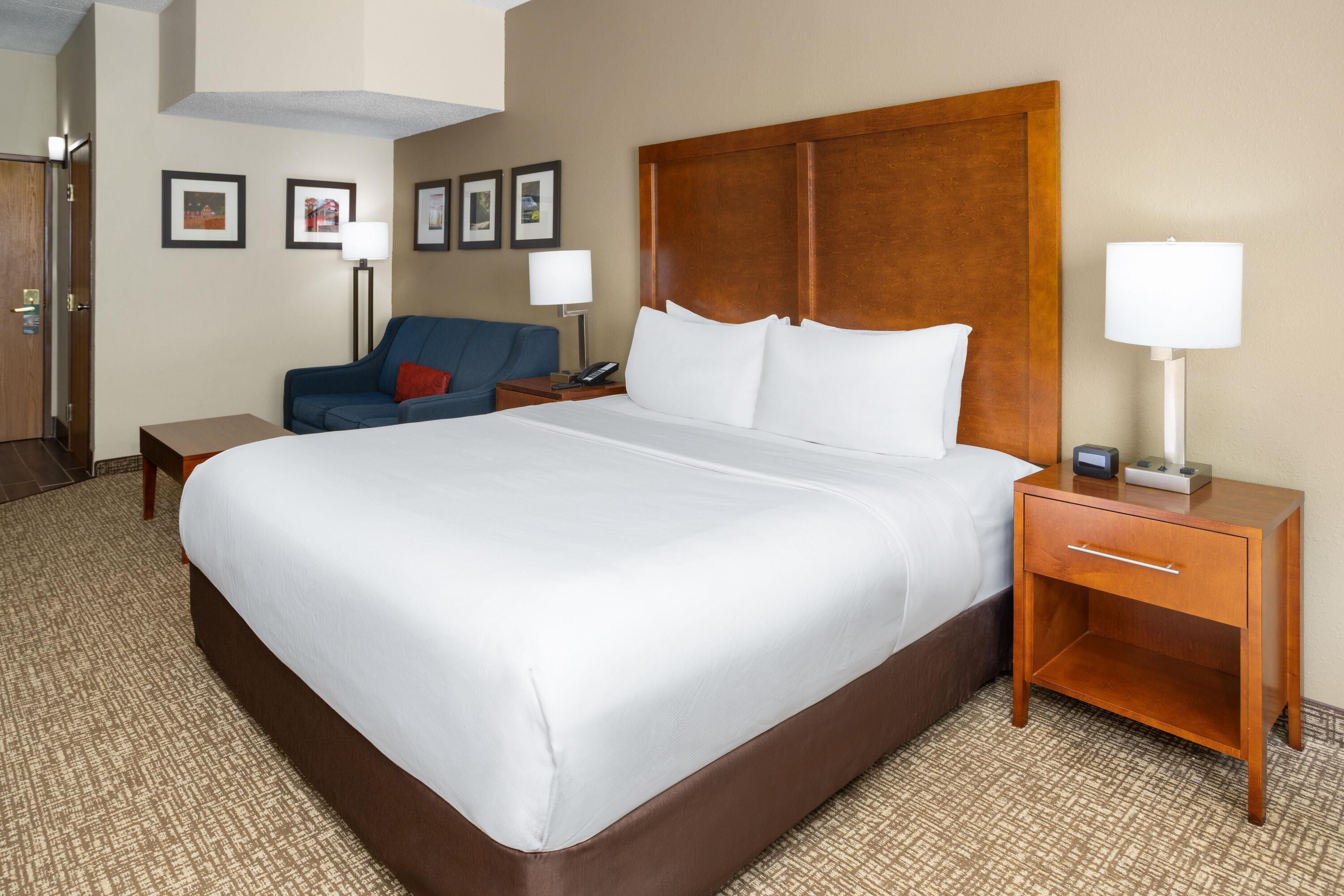 A hotel room with a large bed, a bedside table with a lamp, a chair, and a desk with a phone.