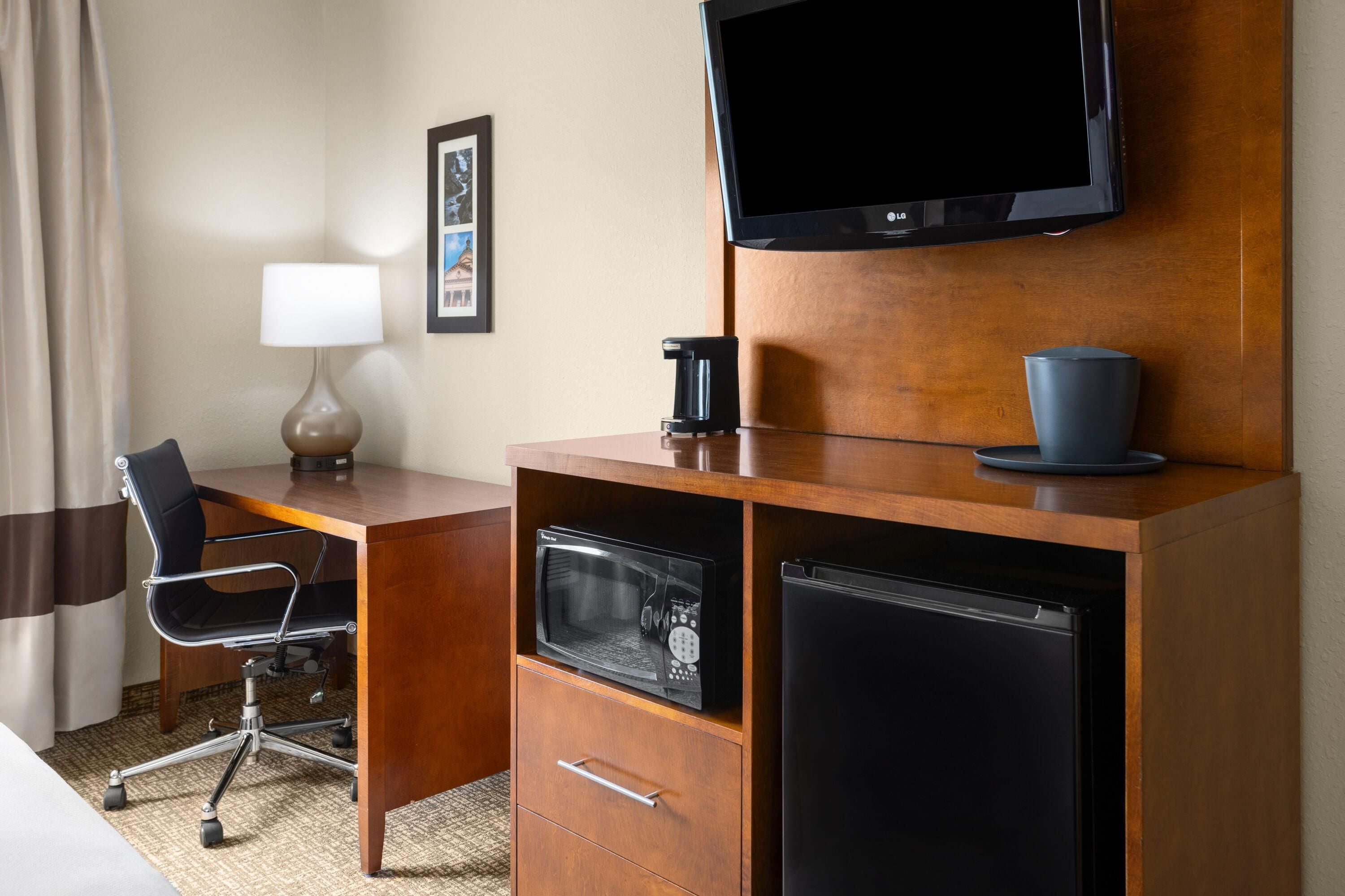 A hotel room with a wooden desk, a television mounted on the wall, a microwave, a refrigerator, a chair, a lamp, and a framed picture on the wall.