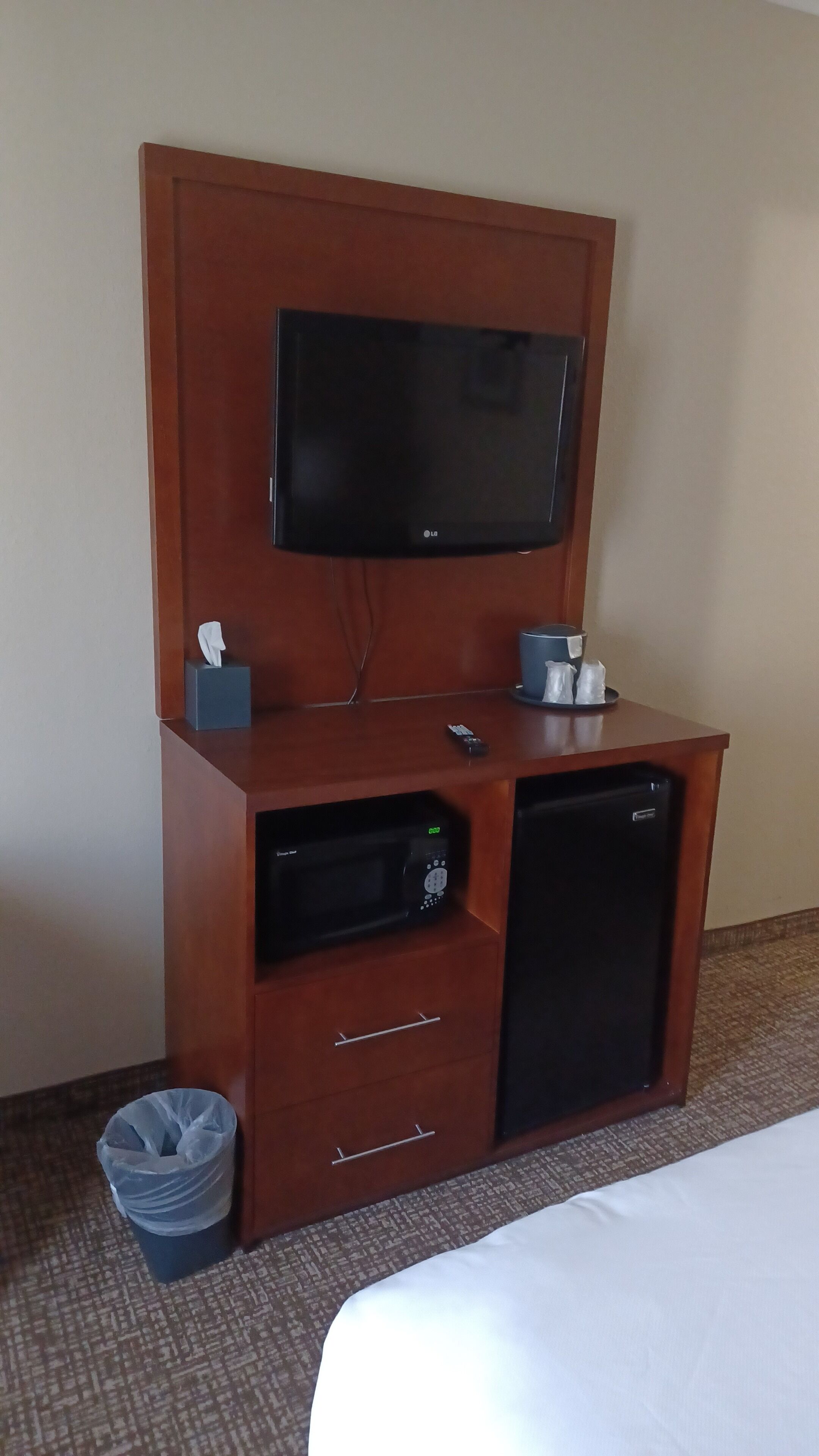 A hotel room with a wooden desk, a flat-screen TV, a mini-fridge, a trash bin, and a bed.