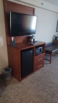 A compact hotel room with a flat-screen TV mounted on a wooden cabinet, a small refrigerator, a microwave, and a trash bin.