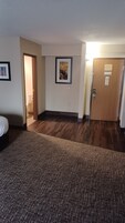 A hotel room with a carpeted floor, a wooden door, a framed picture on the wall, and a visible bathroom.