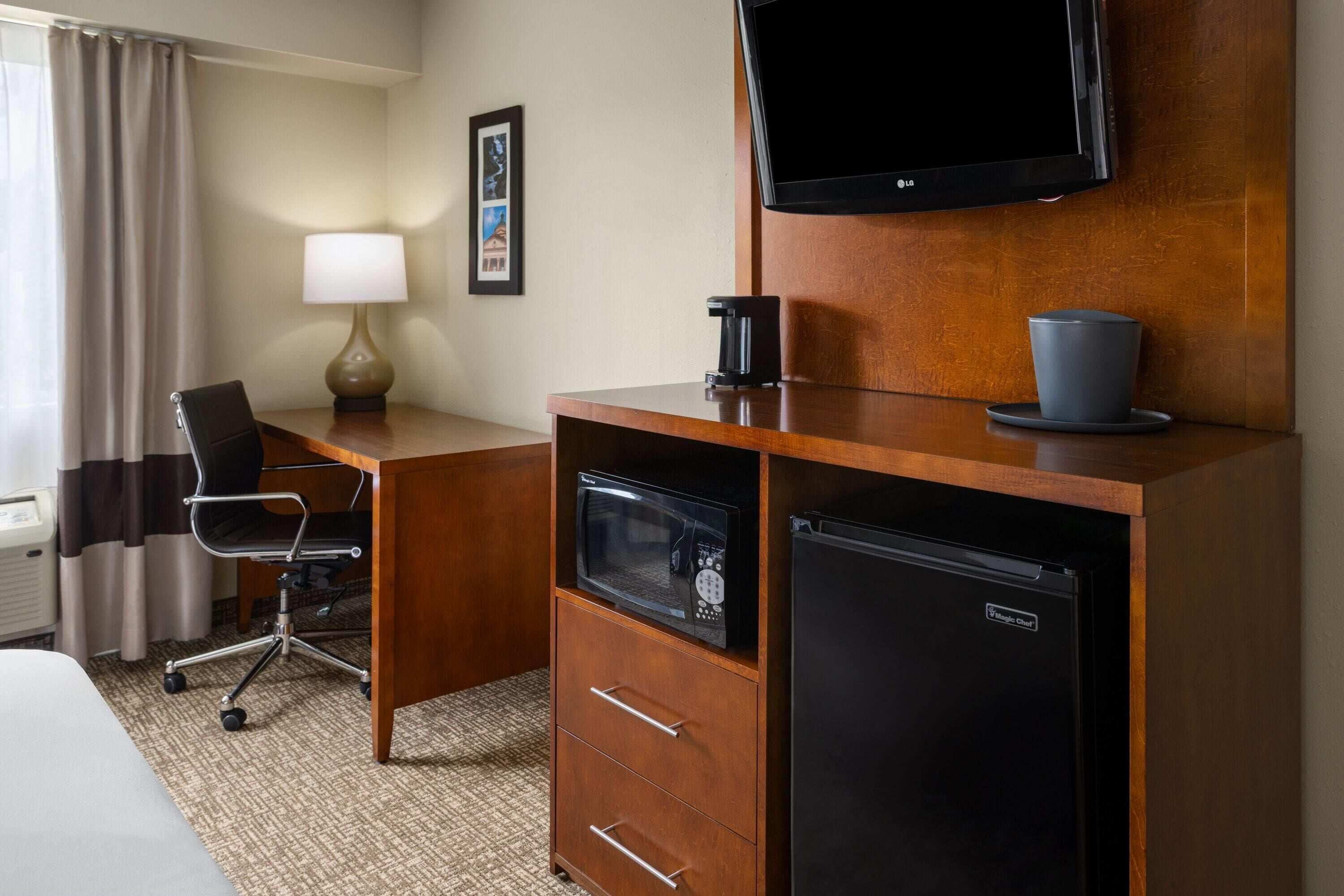 Quarto de hotel com escrivaninha de madeira, frigorífico preto, micro-ondas, máquina de café, televisão, cadeira e cama.