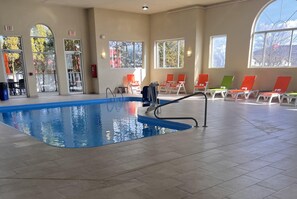 An indoor swimming pool with clear glass walls, multiple exits, and sunlit seating areas.