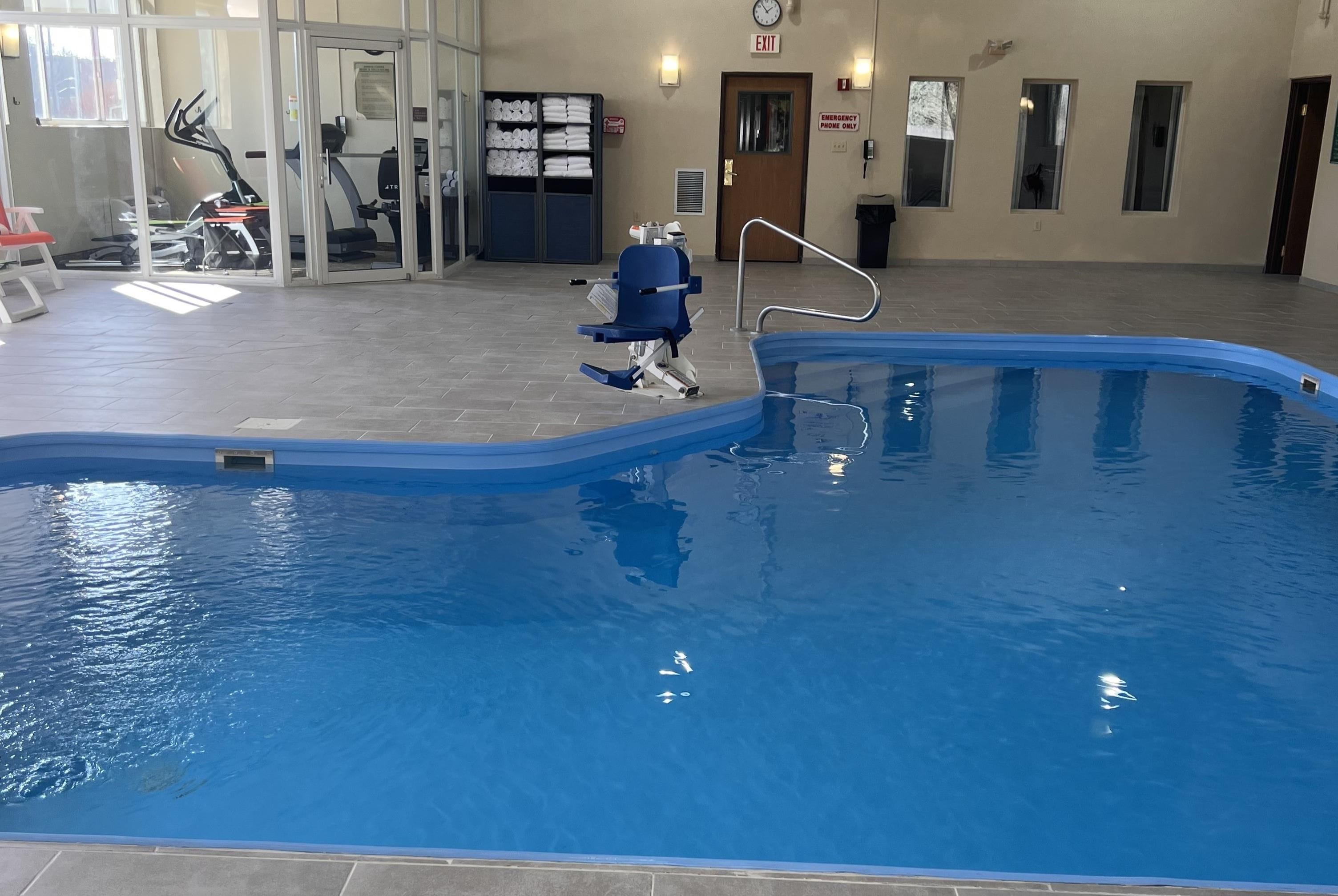 An indoor swimming pool with a blue color, a door with an exit sign, and exercise equipment in the background.