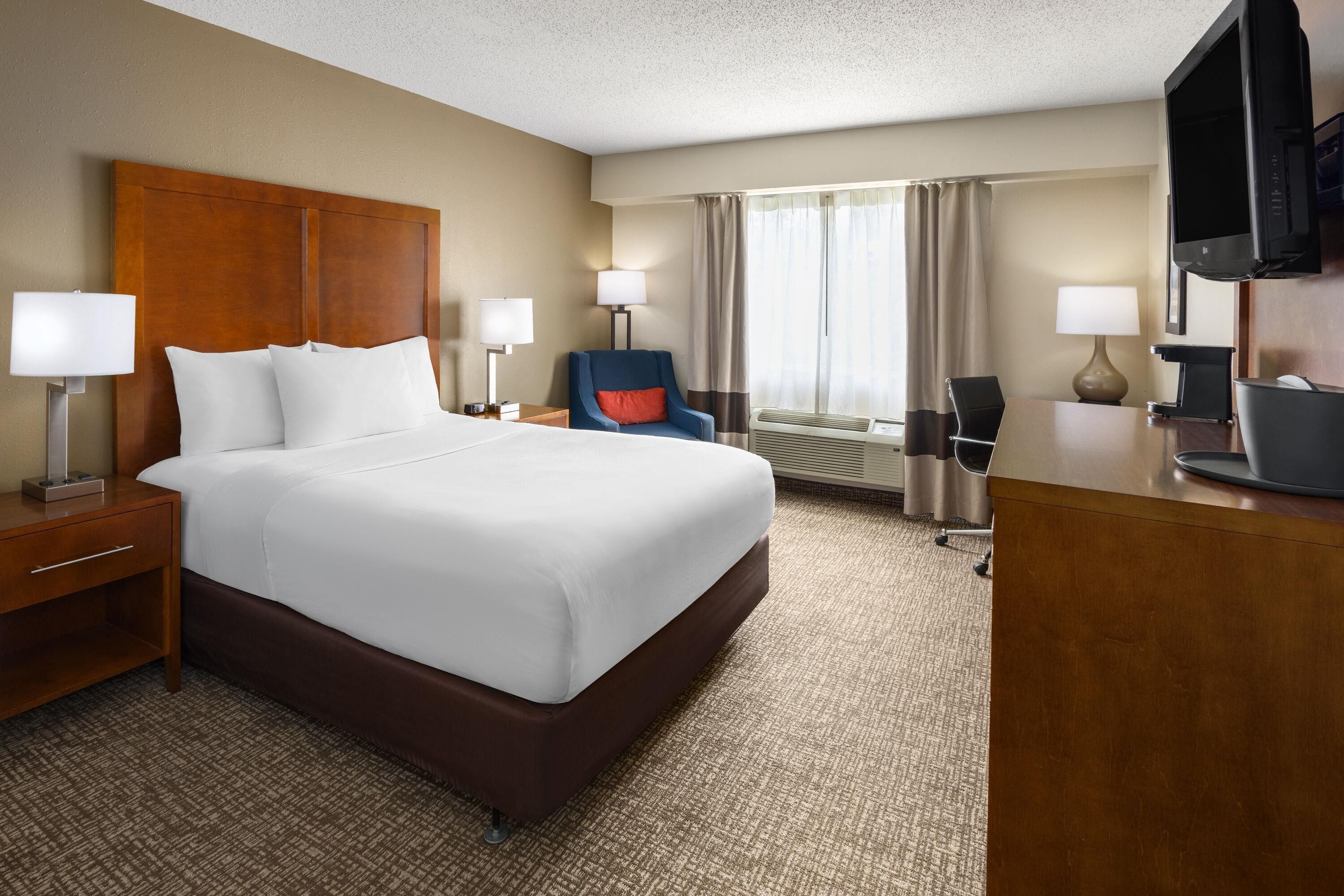 A hotel room with a large bed, two bedside tables with lamps, a desk with a chair, a TV, and a window with curtains.