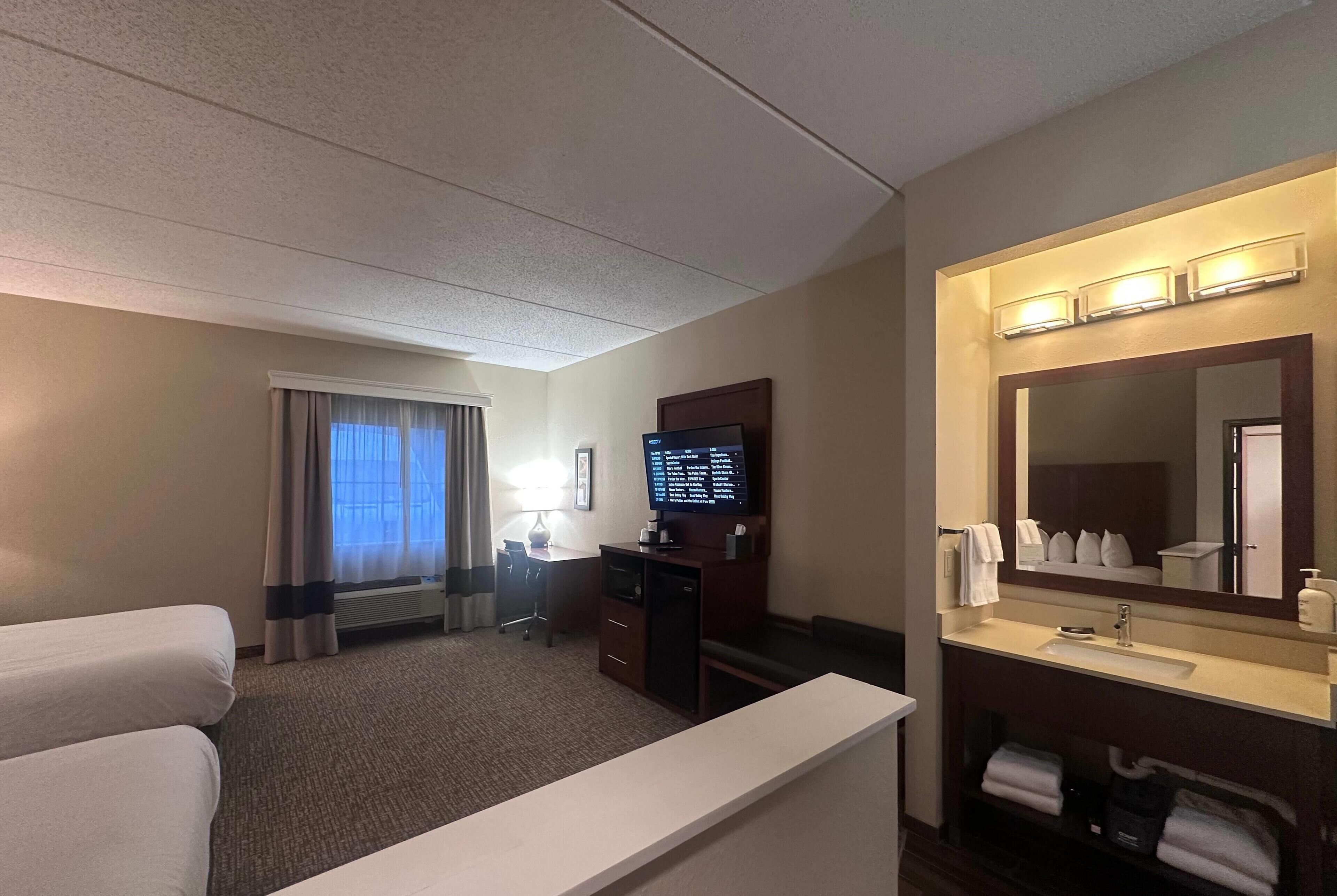 Ein Hotelzimmer mit Bett, Schreibtisch mit Computer, Badezimmer mit Waschbecken und Spiegel sowie Fenster mit Vorhängen.