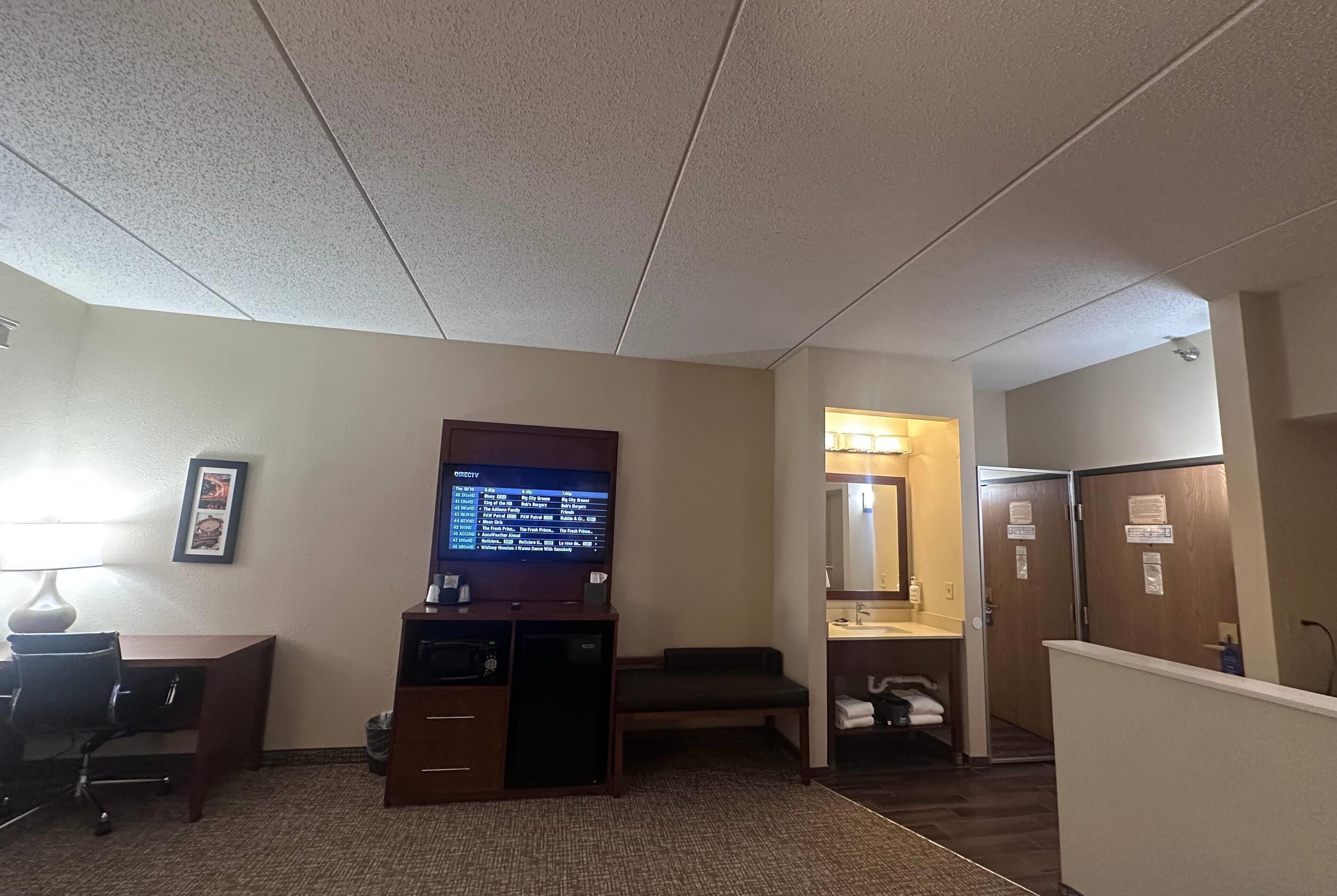 Habitación de hotel con escritorio, televisión, sofá y baño con lavamanos y espejo.