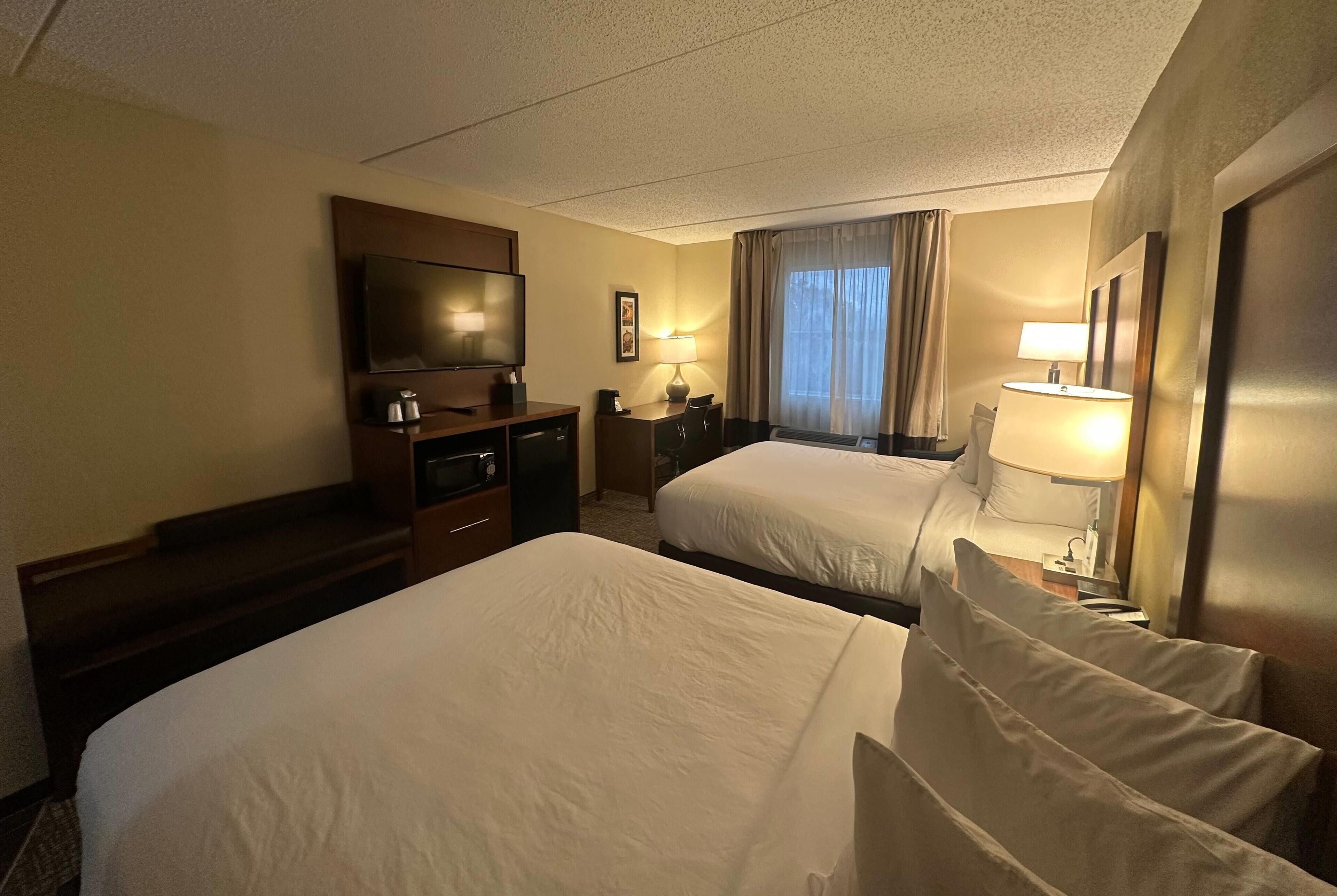 A hotel room with two beds, a TV, a desk with a lamp, and a window with curtains.