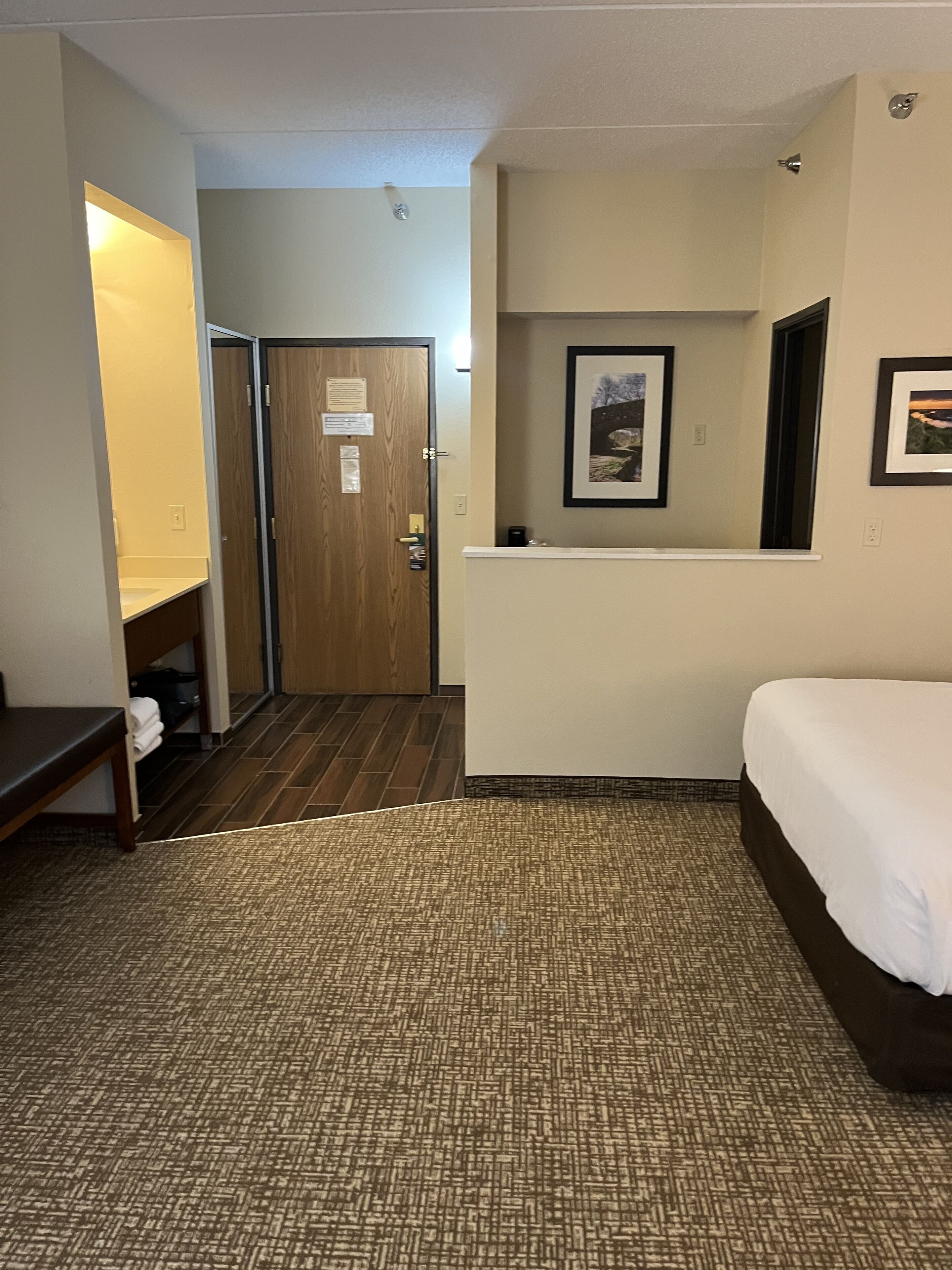 Habitación de hotel con cama, escritorio, silla, mesita con lámpara, puerta y un cuadro en la pared.