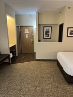 Habitación de hotel con cama, escritorio, silla, mesita con lámpara, puerta y un cuadro en la pared.