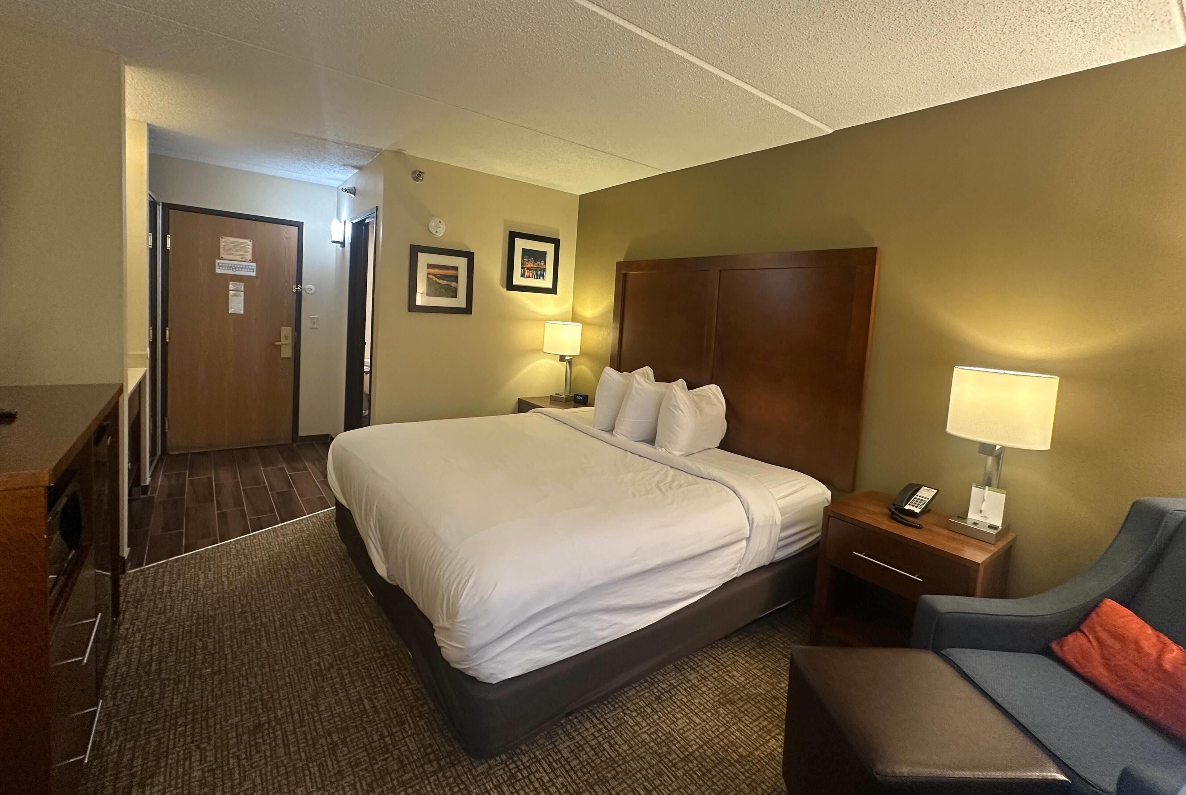 A hotel room with a large bed, a desk with a lamp, a chair, and a small table with a phone.