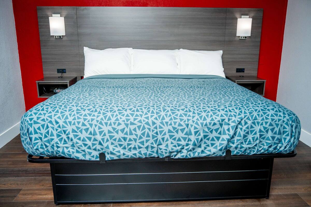 A bed with a geometric patterned bedspread, a wooden headboard, bedside lamps, and a nightstand with a power outlet.