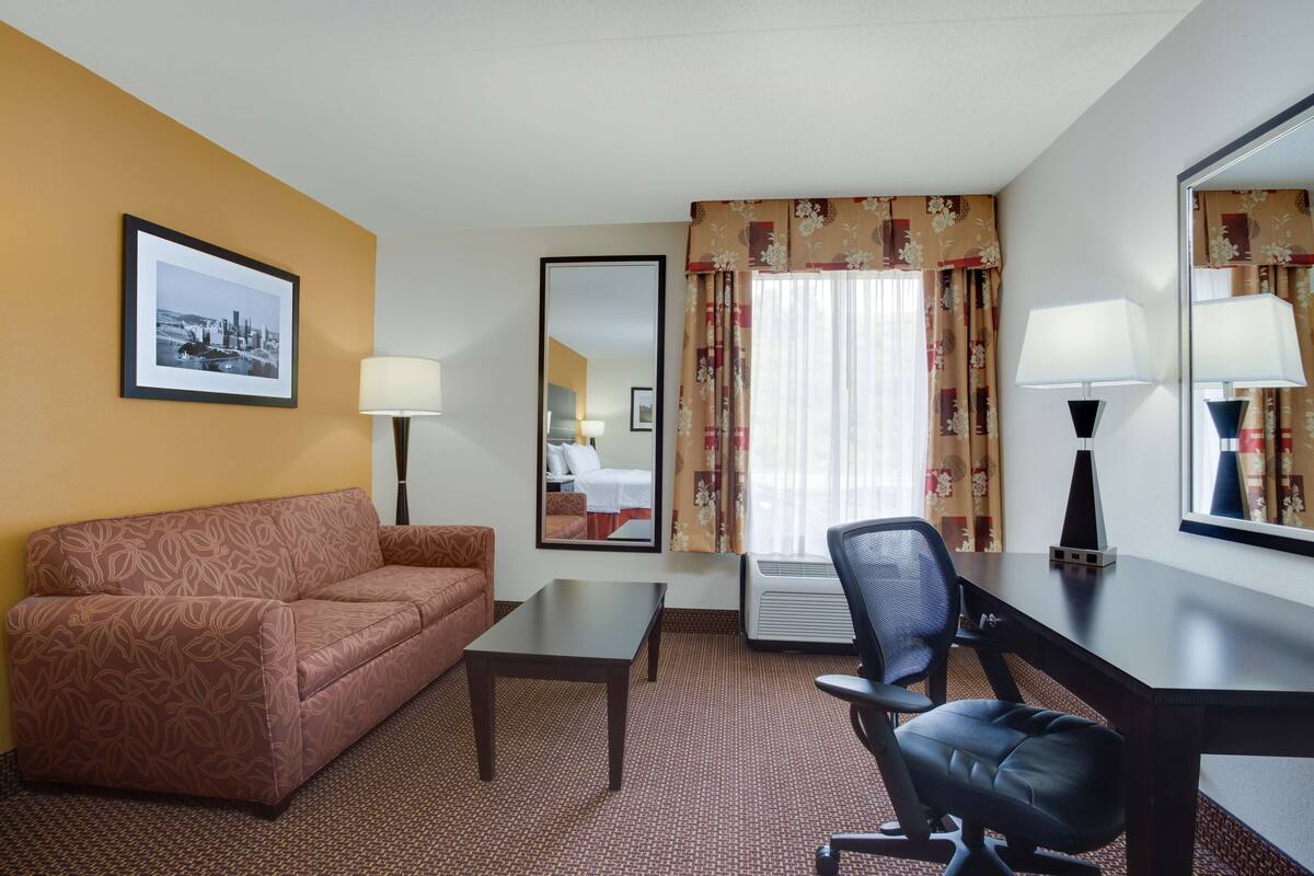 A hotel room with a brown patterned sofa, a small table, a black office chair, a desk with a lamp, a large mirror, and a framed cityscape picture on the wall.