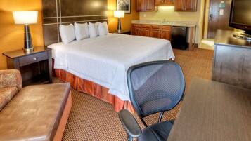 A hotel room with a large bed, a desk, a chair, a television, a desk lamp, a nightstand, a sofa, and a kitchen area with cabinets and a refrigerator.