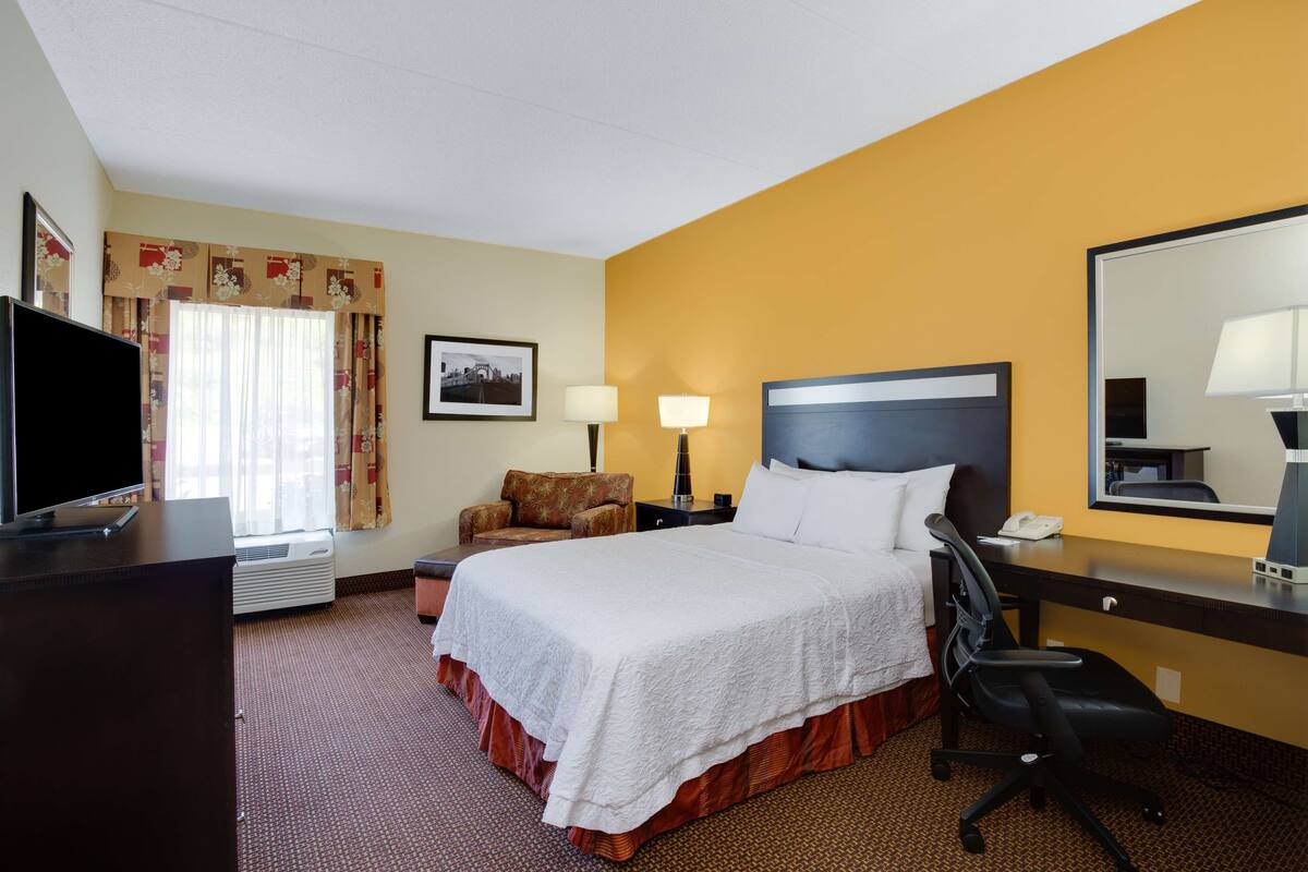 A hotel room with a large bed, a desk with a chair, a television, and a window with curtains.