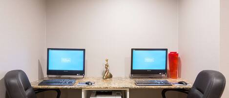 A hotel room with two desks, each with a computer and keyboard, and a printer on a shelf.