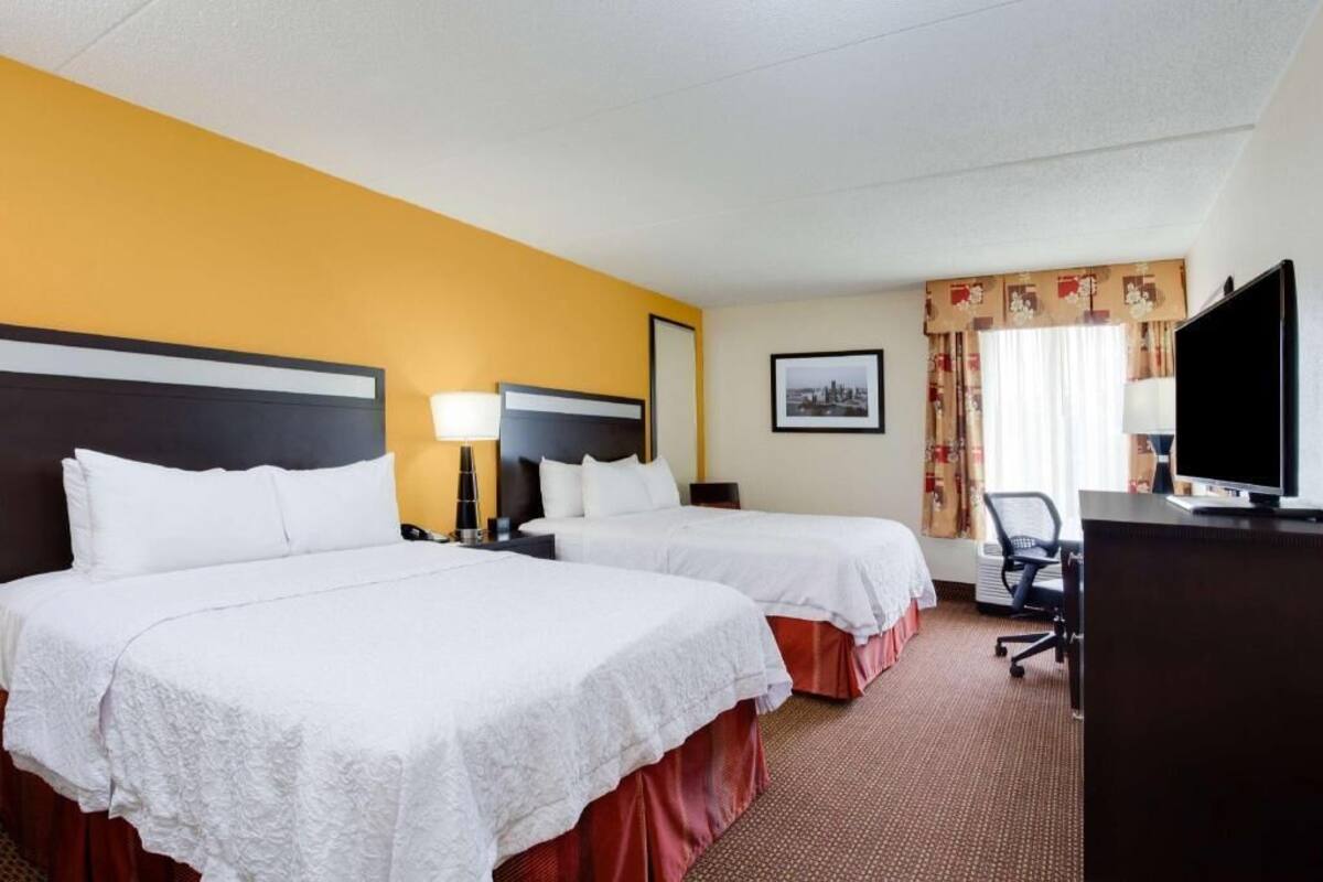A hotel room with two beds, a desk, a TV, and a window with curtains.