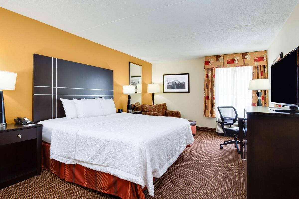 A hotel room with a large bed, two bedside tables with lamps, a desk with a chair, a television, and a window with curtains.