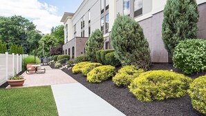 Un edificio moderno con un sendero pavimentado, setos cuidados y una zona de estar con sillas y una mesa.