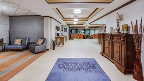 Un hall de hotel con mostrador de recepción, zona de estar con sofás y sillones, una alfombra decorativa con un mensaje motivador y una lámpara de araña grande.