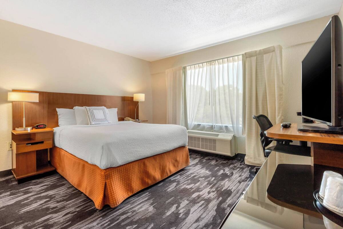 A hotel room with a bed, bedside table, lamp, TV, desk, chair, and window with curtains.